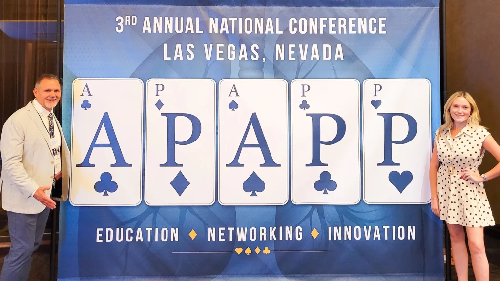 Bryan Miller and Abby Cohen stand on either side of a large banner for the 3rd Annual National Conference in Las Vegas, Nevada, featuring oversized playing cards spelling "APAPP" and the words Education, Networking, Innovation.
